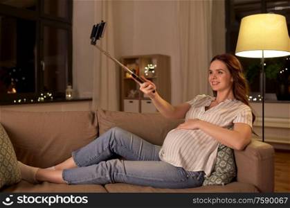 pregnancy, technology and people concept - happy smiling pregnant woman taking picture with smartphone on selfie stick on sofa at home. happy smiling pregnant woman taking selfie at home