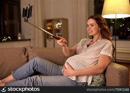 pregnancy, technology and people concept - happy smiling pregnant woman taking picture with smartphone on selfie stick on sofa at home. happy smiling pregnant woman taking selfie at home