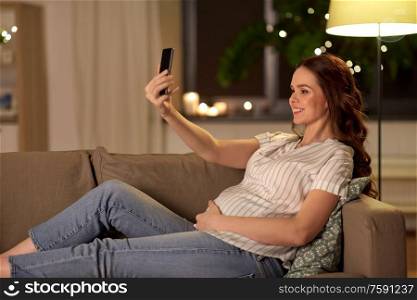 pregnancy, technology and people concept - happy smiling pregnant woman taking selfie with smartphone on sofa at home. happy smiling pregnant woman taking selfie at home