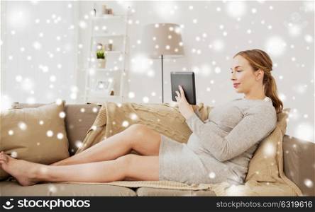 pregnancy, technology and people concept - smiling pregnant woman sitting on couch with tablet pc computer at home over snow. smiling woman with tablet pc at home