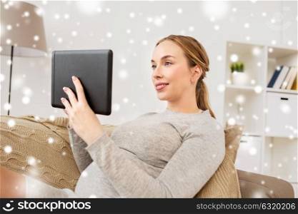 pregnancy, technology and people concept - smiling pregnant woman sitting on couch with tablet pc computer at home over snow. smiling woman with tablet pc at home