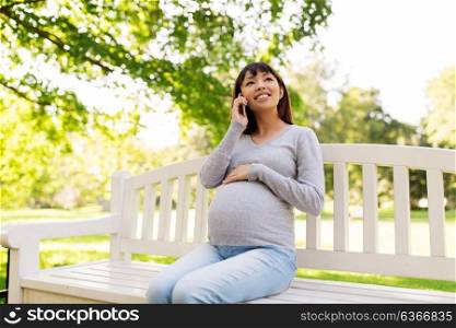 pregnancy, technology, people and communication concept - happy pregnant asian woman calling on smartphone at park. pregnant asian woman calling on smartphone at park