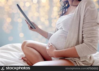 pregnancy, technology, people and expectation concept - close up of pregnant woman with tablet pc computer in bed over holidays lights background