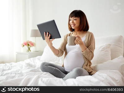 pregnancy, technology, people and expectation concept - happy pregnant asian woman with tablet pc computer drinking tea in bed at home. happy pregnant asian woman with tablet pc at home