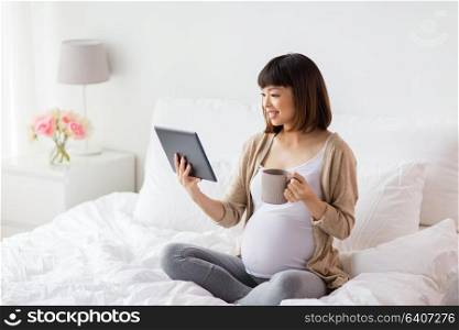 pregnancy, technology, people and expectation concept - happy pregnant asian woman with tablet pc computer drinking tea in bed at home. happy pregnant asian woman with tablet pc at home