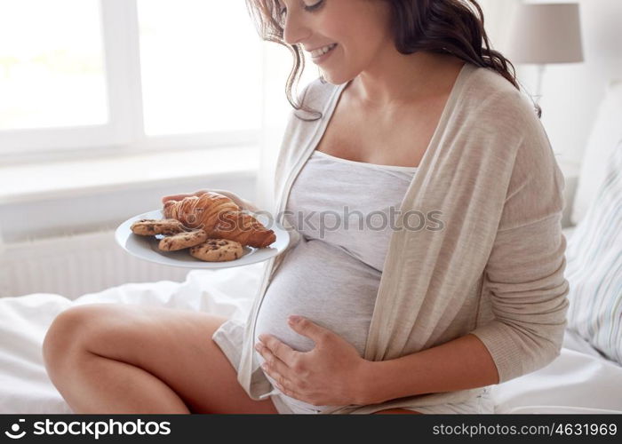 pregnancy, unhealthy eating, food and people concept - close up of happy pregnant woman with croissant buns and cookies sitting in bed at home