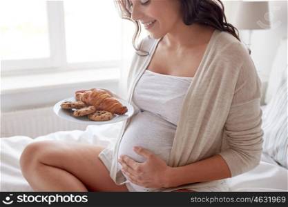 pregnancy, unhealthy eating, food and people concept - close up of happy pregnant woman with croissant buns and cookies sitting in bed at home