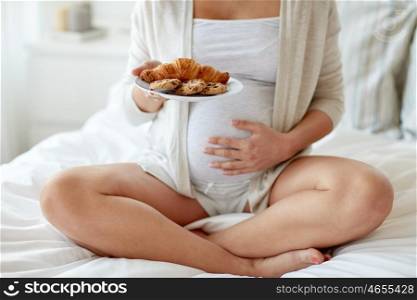 pregnancy, unhealthy eating, food and people concept - close up of pregnant woman with croissant buns and cookies sitting in bed at home