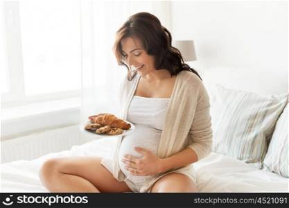 pregnancy, unhealthy eating, food and people concept - happy pregnant woman with croissant buns at home