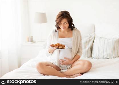 pregnancy, unhealthy eating, food and people concept - happy pregnant woman with croissant buns at home