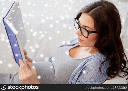 pregnancy, winter and motherhood concept - pregnant woman lying on sofa and reading book at home over snow