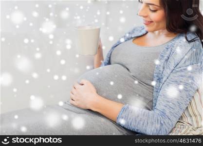 pregnancy, winter, christmas, people and expectation concept - close up of happy pregnant woman with big belly holding cup and drinking tea at home over snow