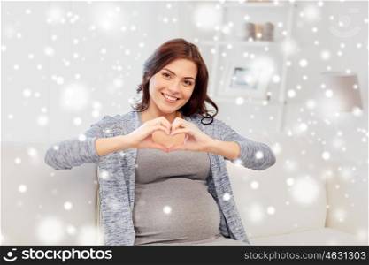 pregnancy, winter, christmas, people and expectation concept - happy pregnant woman sitting on sofa and making heart gesture at home over snow