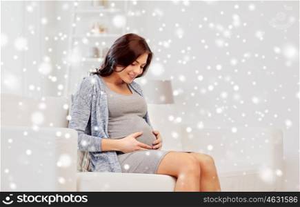 pregnancy, winter, christmas, people and expectation concept - happy pregnant woman sitting on sofa at home over snow