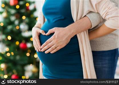 pregnancy, winter holidays and people concept - close up of man making hand heart gesture on his pregnant wife belly at christmas. close up of man and pregnant woman at christmas