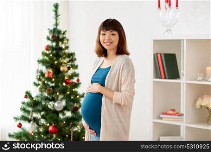 pregnancy, winter holidays and people concept - happy asian pregnant woman at christmas tree at home. happy pregnant woman at christmas tree at home