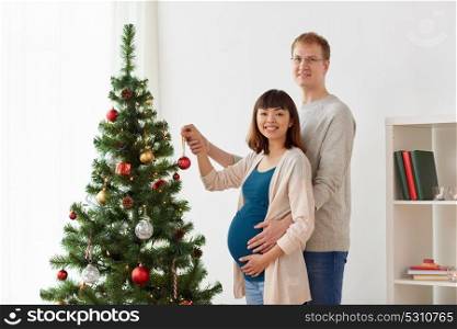 pregnancy, winter holidays and people concept - happy couple decorating christmas tree at home. happy pregnant wife and husband at christmas tree