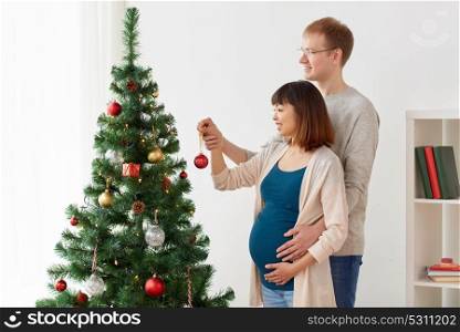pregnancy, winter holidays and people concept - happy couple decorating christmas tree at home. happy pregnant wife and husband at christmas tree