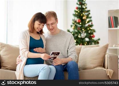 pregnancy, winter holidays and people concept - happy husband and his pregnant wife with baby ultrasound images home at christmas. happy couple with ultrasound images at christmas
