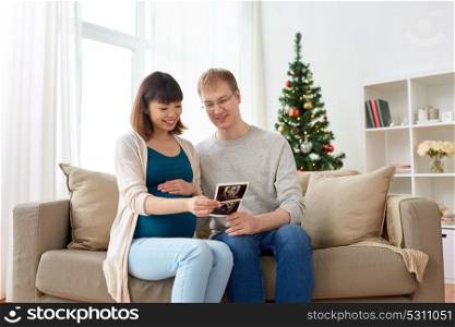 pregnancy, winter holidays and people concept - happy husband and his pregnant wife with baby ultrasound images home at christmas. happy couple with ultrasound images at christmas