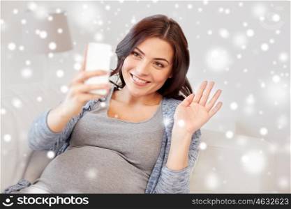 pregnancy, winter, technology, people and expectation concept - happy pregnant woman with smartphone having video call and waving hand at home over snow