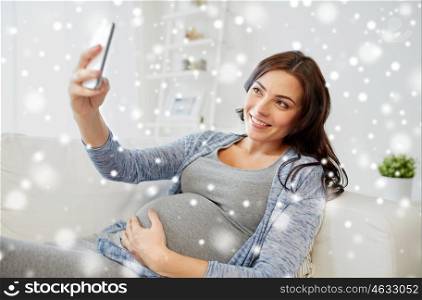 pregnancy, winter, technology, people and expectation concept - happy pregnant woman with smartphone taking selfie at home over snow