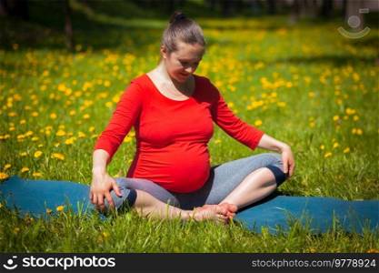 Pregnancy yoga exercise - pregnant woman doing asana baddha konasana bound angle pose outdoors on grass lawn with dandelions in summer. Pregnant woman doing asana baddha konasana outdoors