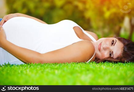 Pregnant arabic woman lying down on fresh green grass field, touching her belly, summer holidays, happy pregnancy concept