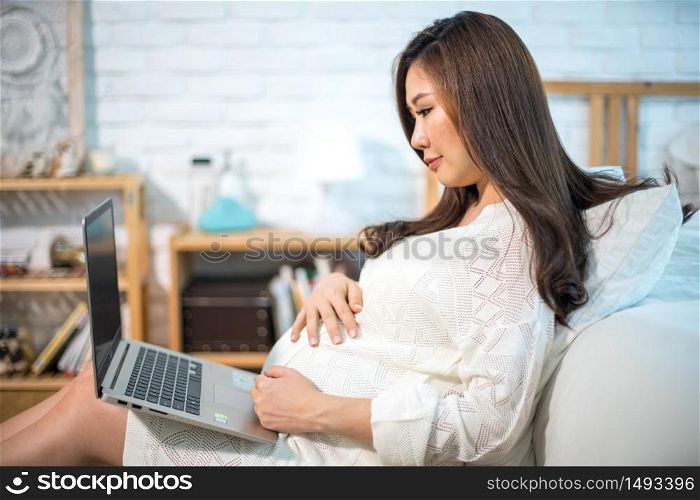 Pregnant asian woman using laptop, sitting on bed at home
