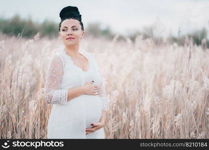 Pregnant beautiful woman in white lace boho dress standing in reeds and holding her belly.. Pregnant beautiful woman in white lace boho dress standing in reeds and holding her belly