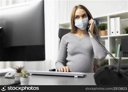 pregnant businesswoman phone her office desk while wearing medical mask