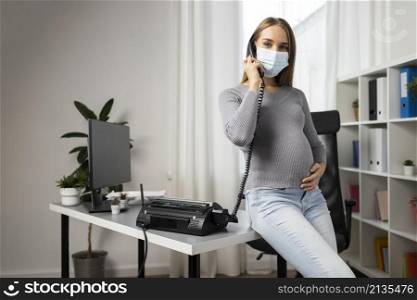pregnant businesswoman taking calls office while wearing medical mask