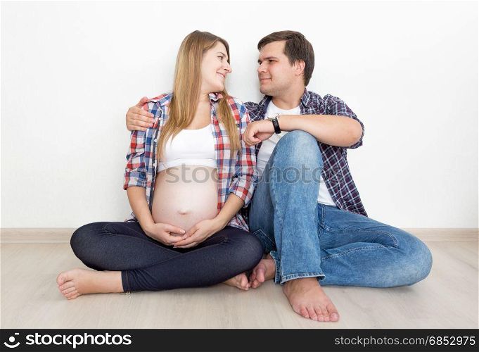 Pregnant couple in jeans and shirts sitting on floor at empty room