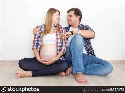 Pregnant couple in jeans and shirts sitting on floor at empty room