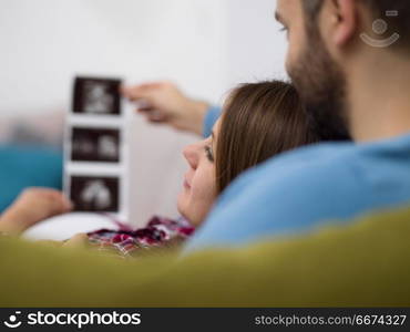 pregnant couple looking baby&rsquo;s ultrasound. Young pregnant couple looking baby&rsquo;s ultrasound photo while relaxing on sofa at home