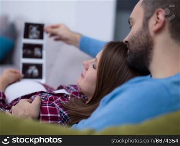 pregnant couple looking baby&rsquo;s ultrasound. Young pregnant couple looking baby&rsquo;s ultrasound photo while relaxing on sofa at home