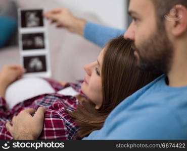 pregnant couple looking baby&rsquo;s ultrasound. Young pregnant couple looking baby&rsquo;s ultrasound photo while relaxing on sofa at home