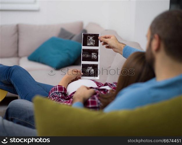 pregnant couple looking baby&rsquo;s ultrasound. Young pregnant couple looking baby&rsquo;s ultrasound photo while relaxing on sofa at home