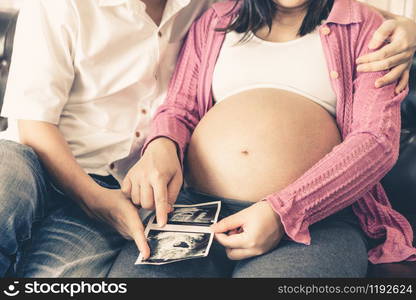 Pregnant couple of husband and wife feels love and relax at home. Young expecting woman holds baby in pregnant belly. Father take care of pregnant mother. Concept of maternity and pregnancy care.