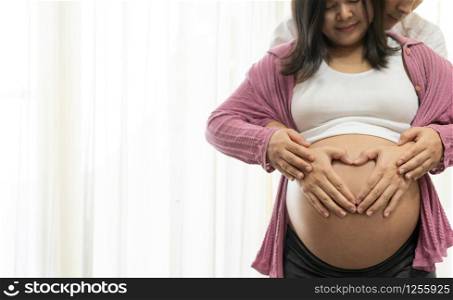 Pregnant couple of husband and wife feels love and relax at home. Young expecting woman holds baby in pregnant belly. Father take care of pregnant mother. Concept of maternity and pregnancy care.