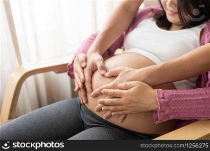 Pregnant couple of husband and wife feels love and relax at home. Young expecting woman holds baby in pregnant belly. Father take care of pregnant mother. Concept of maternity and pregnancy care.