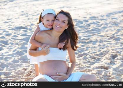 Pregnant mother and daughter on the beach together hug sitting on sand