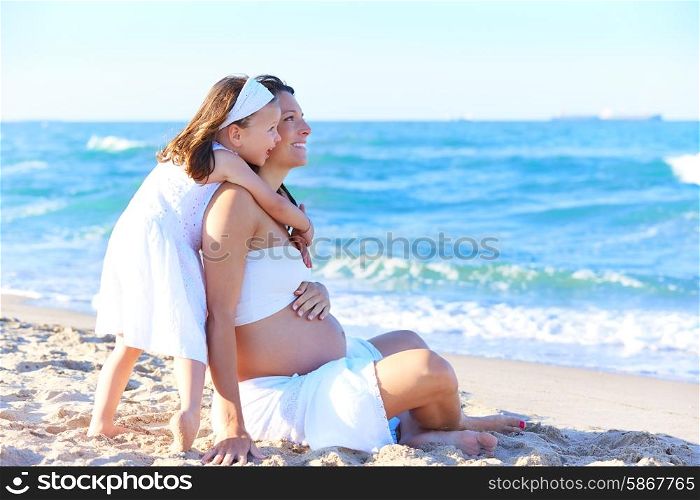 Pregnant mother and daughter on the beach together hug sitting on sand