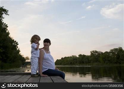 Pregnant Mother And Daughter Sitting On Jetty In Evening
