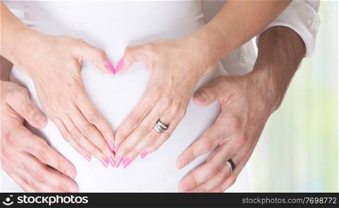 Pregnant woman and her husband holding hands on tummy in heart shape, young loving family, new life concept background