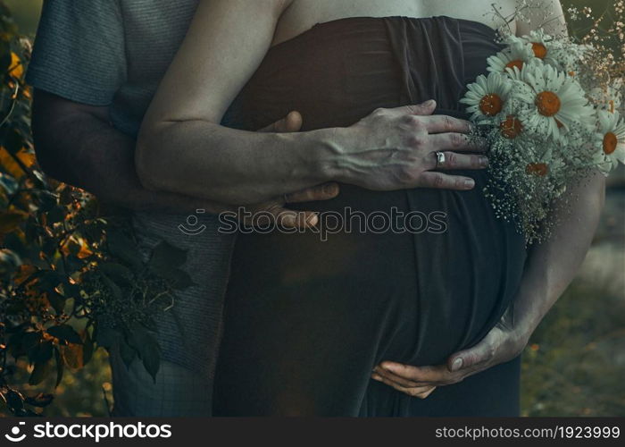 Pregnant woman and her husband hugging her tummy standing outdoors surrounded by nature. Pregnancy, expectation, motherhood concept
