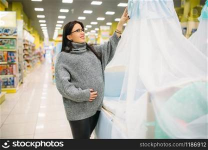Pregnant woman choosing baby cot in store. Future mother in shop of goods for newburns. Pregnant woman choosing baby cot in store