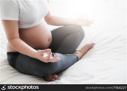 Pregnant woman doing yoga exercise on bed in bedroom at home while taking care of her child. The happy young expecting mother holding baby in pregnant belly. Maternity prenatal care and pregnancy.