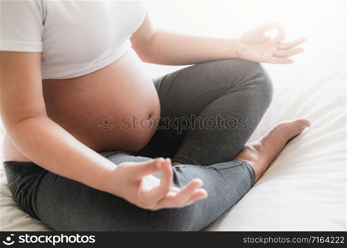 Pregnant woman doing yoga exercise on bed in bedroom at home while taking care of her child. The happy young expecting mother holding baby in pregnant belly. Maternity prenatal care and pregnancy.