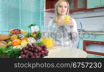 Pregnant woman drinking orange juice in the kitchen near a lot of vegetables and fruits